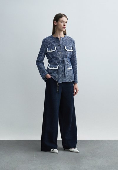 Γυναίκα που στέκεται φορώντας ένα μπλε υφασμάτινο σακάκι με ζώνη και άσπρες τσέπες με περιγράμματα, φαρδιά σκούρα παντελόνια και άσπρα μυτερά παπούτσια.