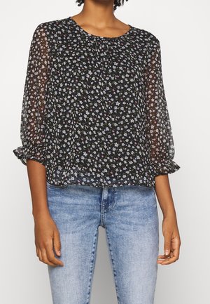 Vrouw draagt een zwarte doorzichtige blouse met bloemenprint, driekwart pofmouwen en lichtblauwe spijkerbroek.