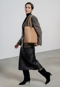 Tan leren tote bag met een gladde textuur, rechthoekige vorm en lange handvatten, gecombineerd met een grijze blazer en een zwarte satijnen rok.