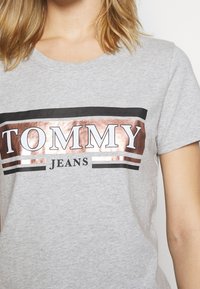 Žena v světle šedém tričku s krátkými rukávy, které má lesklé růžově zlaté a černé vodorovné pruhy a text "TOMMY JEANS" na přední straně.