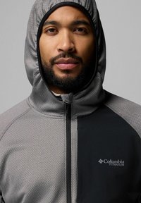 Szara bluza z kapturem o teksturowanym wzorze, z czarnym panelem akcentującym, zamkiem błyskawicznym z przodu i logo Columbia Titanium. Dopasowany krój z kapturem.