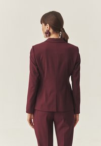 Bordeaux blazer met een gestructureerde silhuet, enkele achteropening en een gladde textuur. Gekleed met bijpassende broek.
