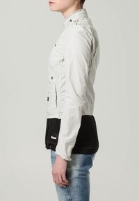 Jasnoszara, krótka kurtka z kołnierzem stojącym, boczną kieszenią i detalami z guzikami. Nosi na sobie czarną bluzkę i zestawiona z wyblakłymi, niebieskimi dżinsami.