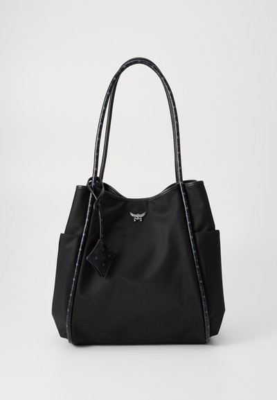 Saco tote preto com duas alças longas com padrão, bolsos laterais, uma pequena etiqueta pendurada e um emblema prateado em forma de asa na frente.