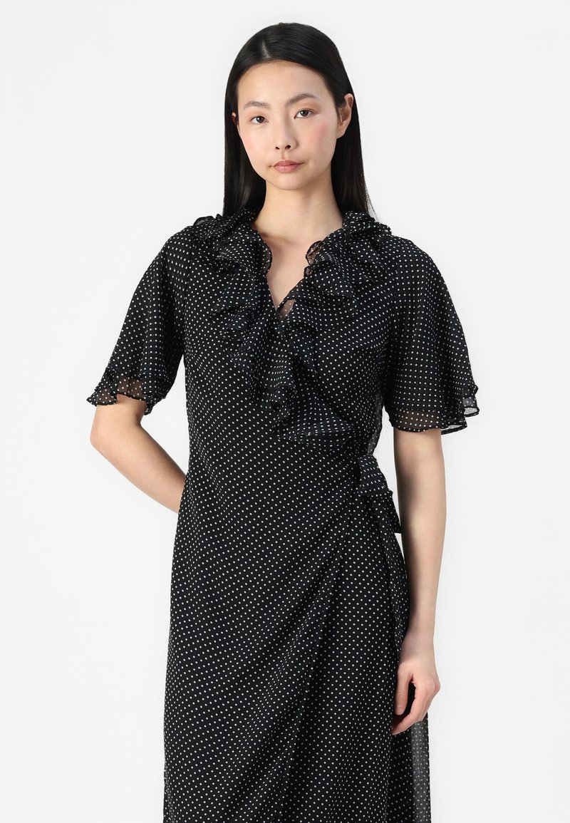 Robe noire à pois blancs, avec un col à volants, des manches courtes et un design croisé. Fabriquée en tissu léger avec une texture douce.