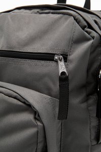 Sac à dos en tissu gris avec plusieurs poches zippées, des coutures solides et des bretelles réglables. Matériau texturé avec des accents subtils.