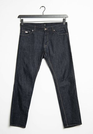 Tmavě modré džíny s pěti kapsami zavěšené na černé ramínko na bílém pozadí.