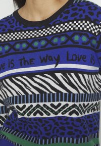 Pull avec des motifs bleus, noirs, blancs et verts comprenant des imprimés léopard, zèbre, des cœurs, et un texte "Love is the way" sur la poitrine.