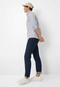 Kariertes Hemd mit kleinen blauen Blumenmustern, lange Ärmel hochgerollt, kombiniert mit dunklen Jeans und weißen Sneakers. Beigefarbene Cap rückwärts getragen.