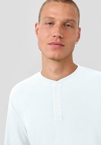 Junger Mann mit kurzen Haaren und Ohrringen, der ein weißes, geknöpftes Henley-Shirt vor einem schlichten hellen Hintergrund trägt.