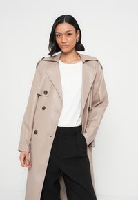 Μπεζ παλτό τύπου καπαρντίνα από δερματίνη, με διπλό πέτο, μεγάλα κουμπιά και επιγουντιαίες επιγονατίδες, συνδυασμένο με λευκό πουκάμισο και μαύρο παντελόνι.