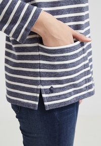 Marinblå och vit randig långärmad topp med ficka fram och sidsplits. Mjuk, texturerad tyg med en liten broderad logotyp.