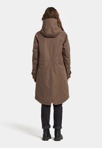 Bruine gewatteerde jas met een licht aflopende ontwerpelement, met een halflange snit en knoopaccenten op de mouwen. Gecombineerd met donkere jeans en laarzen.