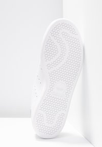 Valkoisen urheilukengän pohja, jossa on teksturoitu mehiläishoitokuvio, kohokuvioitu logo ja sileä, kumipintainen pinta.