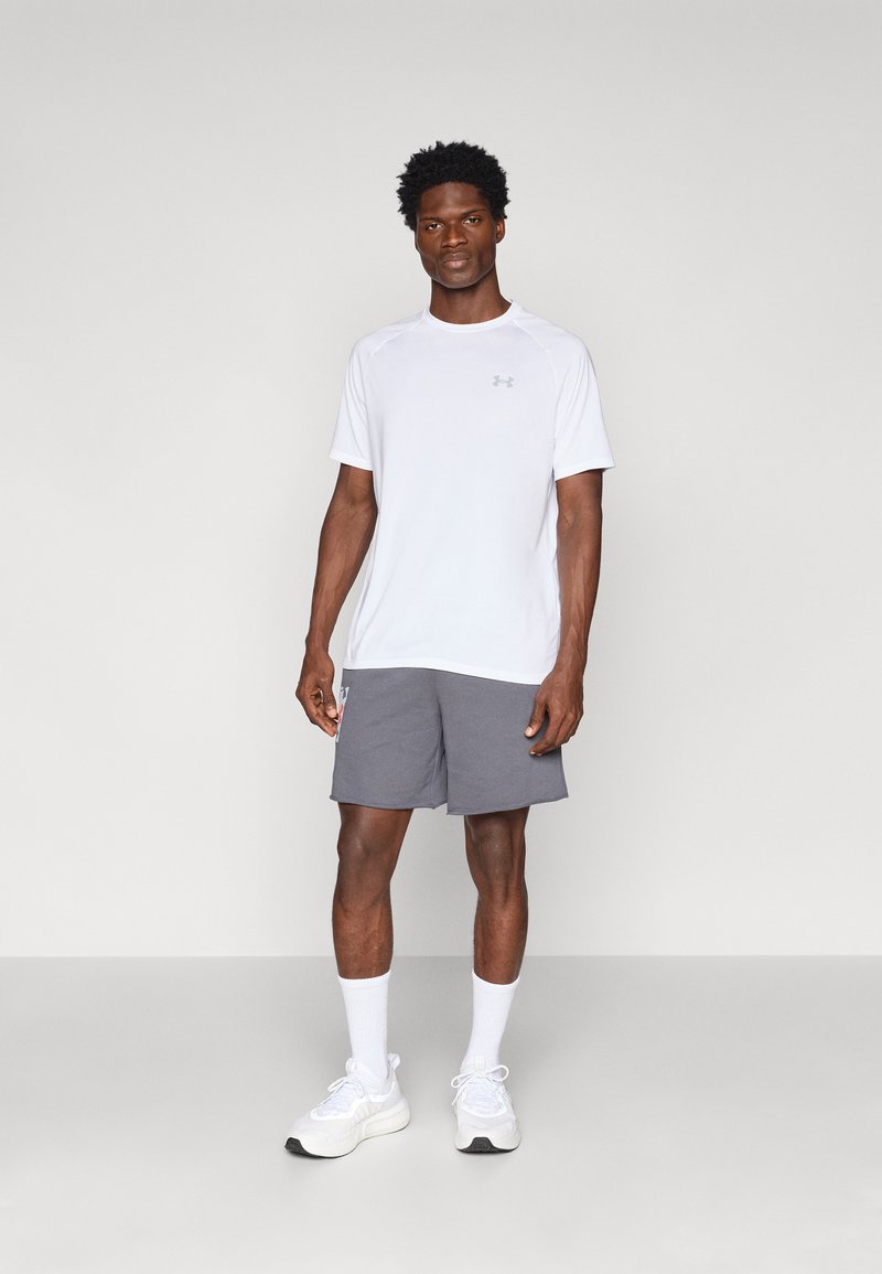 Weißes Sport-T-Shirt und graue Shorts, mit einem Logo auf dem Shirt und den Shorts. Das Modell trägt weißeSneaker und Socken in Knöchellänge.