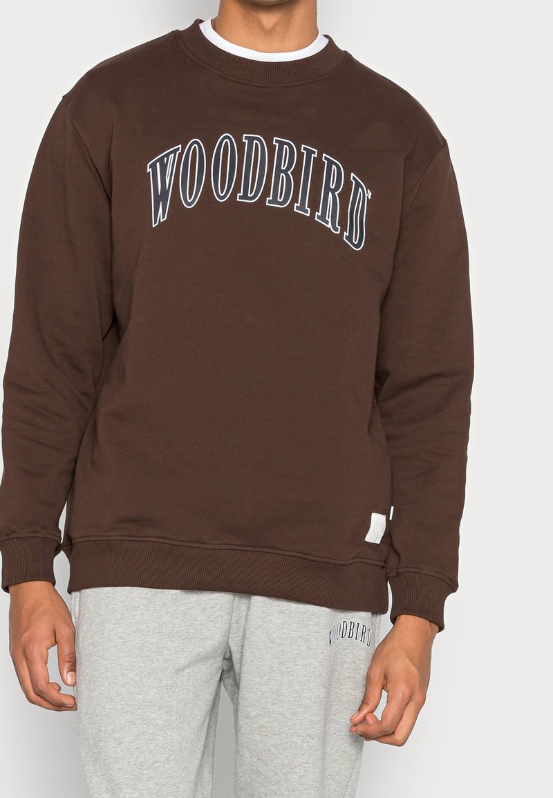 Sudadera marrón con cuello redondo y texto arqueado en blanco que dice "WOODBIRD". Puños y dobladillo acanalados, combinados con pantalones de chándal gris claro.