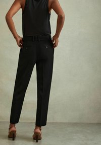 Schwarze, taillierte Hose mit passgenauer Schnittführung, sichtbaren Gürtelschlaufen und zwei Gesäßtaschen mit Knopfverschluss. Kombiniert mit braunen, hohen Sandalen.