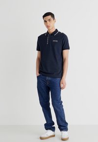 Jūras spēku polo krekls ar baltu apkakles noformējumu, savienots ar ziliem džinsiem. Apģērbs ietver baltos sporta apavus un vienkāršus dizaina elementus.