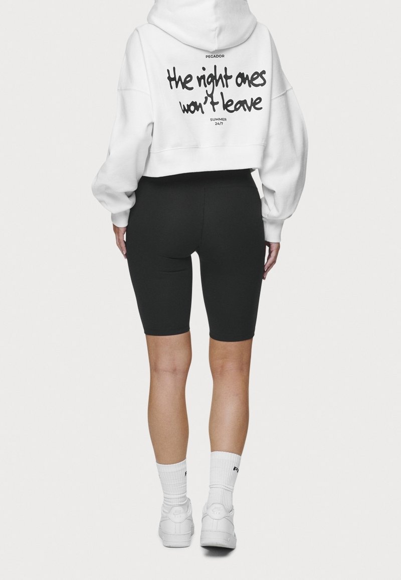 Sudadera blanca corta con texto negro en la parte de atrás y pantalones cortos de ciclismo negros. Calcetas blancas y zapatillas completan el conjunto.