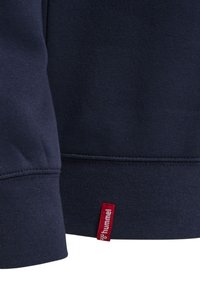 Ciemnoniebieska tkanina z bliska, pokazująca mankiet i szew na dole z małą czerwoną metką z napisem "hummel" przymocowaną do dolnej krawędzi.