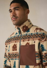 Kurtka polarowa z beżową bazą, brązowymi i turkusowymi wzorami drzew i domów, zapięcie na zamek błyskawiczny oraz brązowa kieszeń na piersi z materiału.