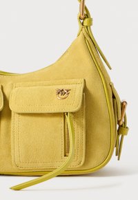 Saco de ombro amarelo em camurça com dois bolsos frontais, detalhe em zipper e acessórios decorativos, apresentando uma forma curvada e alça ajustável.
