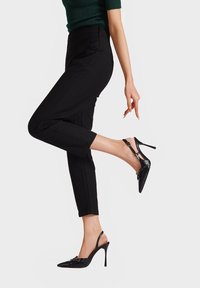 Décolleté neri con punta affusolata, in pelle verniciata lucida, tacco sottile e dettaglio decorativo a fiocco. Abbinati a pantaloni neri aderenti.