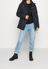 Kobieta w granatowej kurtce z kapturem, czarnym swetrze dzianinowym, jasnoniebieskich spodniach dżinsowych z podwiniętymi nogawkami i czarnych sztywnych butach, stojąca na jednolitym tle.