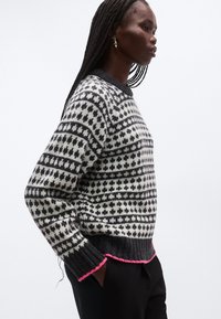 Stickad tröja i svart och gräddvitt med geometriska mönster. Har ribbad nederkant och mudd med rosa accenter. Modellen visas i profil.
