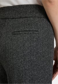 Gros plan sur une personne portant un pantalon à motif chevrons gris foncé avec une poche passepoilée à l'arrière et un bras nu visible.