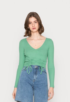 Blusa verde de mangas longas com nervuras, apresentando um decote em V, detalhe de cintura franzida com cordões, combinada com calções de ganga azuis de cintura alta.