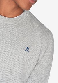 Suéter gris claro con un tejido texturizado, escote redondo y un pequeño logo bordado azul en el pecho.