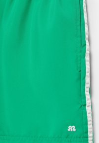 Tissu vert avec bordure latérale blanche et un petit logo blanc brodé près du bord inférieur, probablement une partie d'un vêtement.