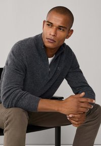 Pull en maille grise avec un col zippé, des poignets côtelés et une coupe décontractée. Le modèle est assis, portant un pantalon marron et une chemise blanche en dessous.