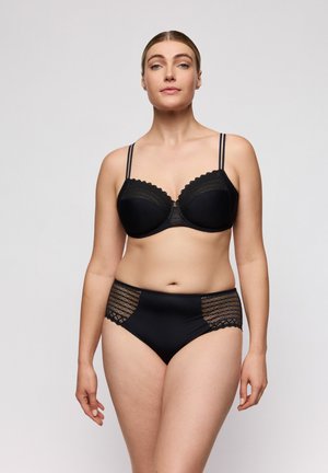 Vrouw staand, gekleed in een zwarte kanten beha met dubbele bandjes en bijpassend hoog getailleerd kanten ondergoed, tegen een effen grijze achtergrond.