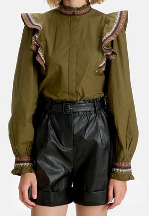 Olijfgroene blouse met ruches op de schouders en geborduurde manchetten, gecombineerd met hooggetailleerde zwarte leren shorts met riem.