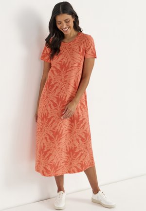 Femme portant une robe midi orange à manches courtes avec des motifs de feuilles et des baskets blanches, debout et souriante contre un mur blanc.
