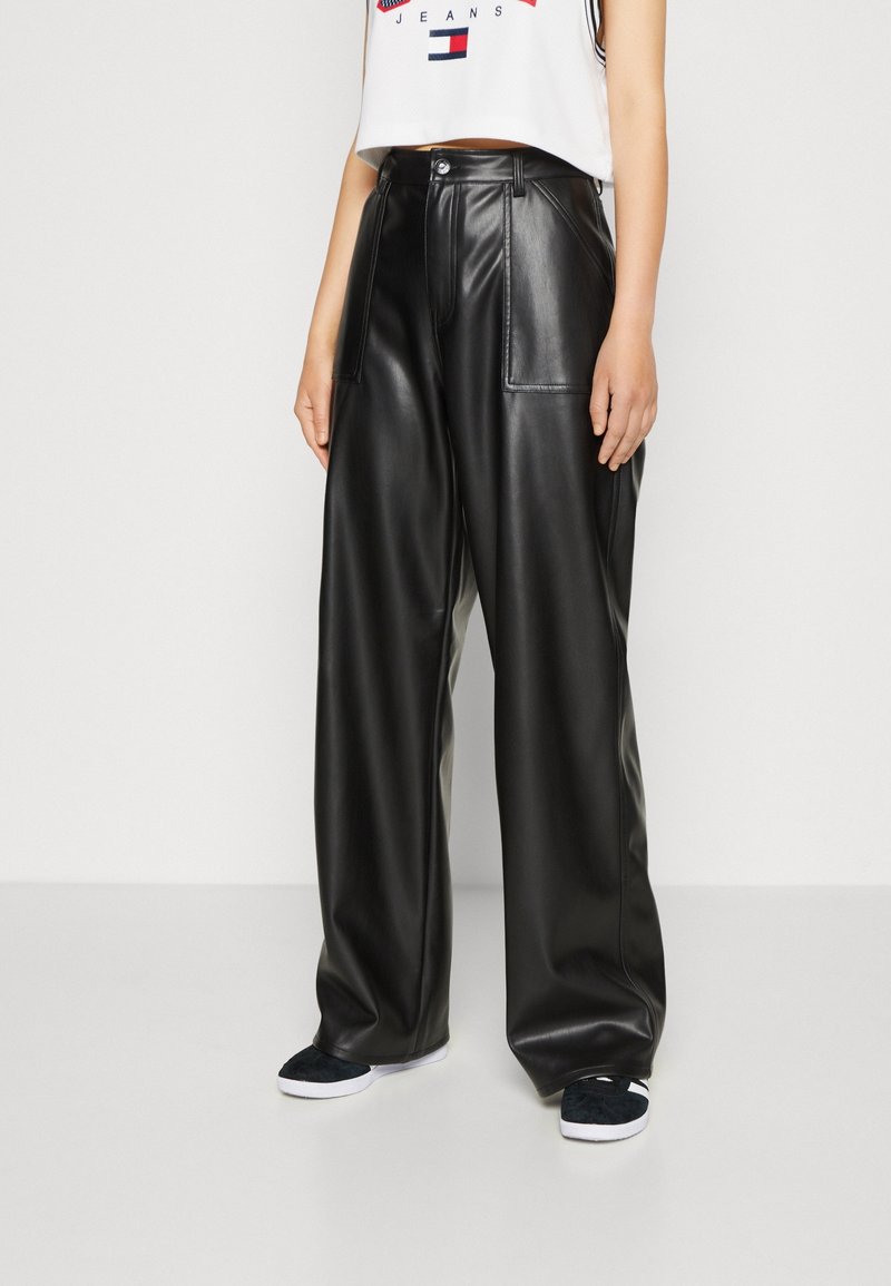 Personne portant un pantalon large en similicuir noir avec de grandes poches avant et des baskets noires à semelles blanches.