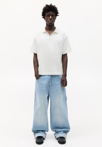Polo alb cu mânecă scurtă; blugi denim supradimensionați de culoare albastru deschis cu un finisaj decolorat; adidași negri; model în picioare cu mâinile în buzunare.