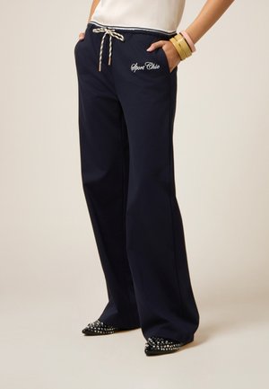 Pantaloni della tuta blu navy con coulisse regolabile a righe, logo ricamato 'Sport Chic' e tasche laterali. Abbinati a scarpe nere con borchie.