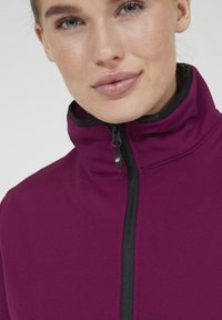 Bordeauxkleurige jack met rits en hoge kraag, met zwarte ritssluiting. De stof lijkt soepel en aansluitend, geschikt voor sportkleding.