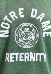 Tissu en mesh vert avec texte blanc usé « MOTRE DAINE » et « RETTERNITY » autour d'un emblème circulaire avec des figures et du texte.