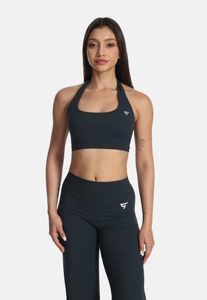 Kobieta stojąca przodem w czarnym sportowym biustonoszu halter i pasujących legginsach z wysokim stanem z logo po lewej stronie na piersi i udzie.
