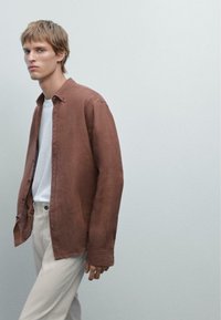 Chemise en lin marron déboutonnée au-dessus d'un T-shirt blanc, accompagnée de pantalons de couleur claire. La chemise a une coupe décontractée et des manches retroussées.