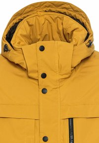 Gul isoleret jakke har en høj krave, lynlås og trykknaplukning samt sorte knapper. Stoffet virker glat med en quiltet inderside.