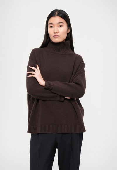 Un pull à col roulé marron avec un tricot texturé, des poignets et un ourlet côtelés. Associé à un pantalon à rayures fines sombres, créant une silhouette décontractée.