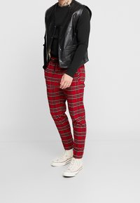 Mężczyzna w czarnej skórzanej kamizelce na czarnej koszuli z długim rękawem, czerwonych spodniach w kratę i białych wysokich trampkach stoi na jednolitym tle.