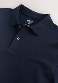 Marineblå poloshirt med krave, to knapper, lille broderet krone-logo og et mærke med størrelsen medium og mærket NEXT.
