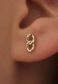 Gouden metalen oorbellen met een verweven schakelontwerp, een gepolijste afwerking en een gladde textuur, bevestigd aan het oorlel van een persoon.