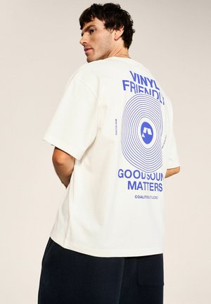Mężczyzna noszący białą, oversize koszulkę z niebieskim, okrągłym wzorem winylowej płyty i napisem "Vinyl Friendly Good Sound Matters" na plecach.